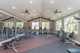 Located in Athens, Georgia, this fitness center offers a well-equipped, single-floor layout designed to support a wide range of workout routines. The space features soaring vaulted ceilings accented by bold burgundy trim, which add architectural character and a sense of openness throughout. Recessed lighting and dual ceiling fans ensure consistent illumination and airflow across the entire floor.

Large white-framed windows line the walls, inviting abundant natural light and offering pleasant views of the surrounding tree canopy. The flooring combines cushioned blue carpet tiles with durable terracotta-toned rubber matting, clearly delineating cardio and strength training zones.

The cardio area is outfitted with multiple treadmills, elliptical trainers, and a stationary cycle. The strength training section includes a cable pulley machine, a leg press/curl station, adjustable benches, and a dumbbell rack. A dedicated functional training zone provides stability balls, resistance bands, and other accessories neatly organized on a rolling storage rack. Full-length mirrors along one wall enhance spatial perception and allow users to monitor form during exercise.

A decorative panel wall featuring a white grid over a deep burgundy background serves as a striking design focal point, while built-in bench storage adds practical convenience. The overall ambiance is clean, energetic, and welcoming.