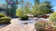 This outdoor space is a thoughtfully landscaped community area that combines functional design with natural greenery. The grounds feature a mix of manicured shrubs, ornamental plantings, and mature trees — including a graceful weeping willow — that provide ample shade and a serene atmosphere throughout the open-air setting.

A prominent gravel garden area serves as a central focal point, bordered by flagstone pathways that guide visitors through the landscape. Neatly trimmed round shrubs and flowering plants add color and structure to the gravel beds, creating a visually balanced and low-maintenance garden design.

A fenced backyard section, enclosed by a tall wooden privacy fence, offers a more intimate outdoor retreat. This area includes a picnic table set on a combination of gravel and flagstone paving, making it a practical space for outdoor dining or gatherings. The fence provides clear boundary definition while a covered structure in the background suggests additional sheltered amenities nearby.

The broader community grounds are framed by mature oak trees and well-kept lawns, with paved walkways connecting various areas. Multi-story residential buildings with brick and neutral-toned facades are visible in the background, reinforcing the communal character of the space. The property enjoys abundant natural light under open skies, contributing to an inviting and well-appointed outdoor environment.