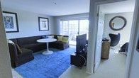 This single-floor space offers a comfortable and well-appointed living environment centered around a thoughtfully designed living room. Large sliding glass doors and wide windows flood the space with natural light, offering views of the surrounding greenery and creating an open, airy atmosphere throughout.

The living room serves as the heart of the home, anchored by a generous dark sectional sofa adorned with colorful accent pillows in blue and green tones. A striking blue distressed area rug adds a bold pop of color and texture to the neutral carpeted flooring beneath. A sleek round white pedestal table provides a modern focal point within the seating area, complementing the contemporary aesthetic. A flat-screen television on a low-profile media stand rounds out the entertainment setup.

Neutral gray walls provide a versatile backdrop that allows the furnishings and décor to stand out, while framed wall art adds a personal touch. An adjacent room, visible through a white paneled doorway, hints at additional living or utility space with warm wood-toned cabinetry. The overall ambiance is clean, modern, and inviting, with a balanced mix of bold accents and understated design elements that make the space feel both stylish and livable.