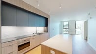 This contemporary single-floor residence showcases a thoughtfully designed open-concept layout with clean lines and a cohesive modern aesthetic throughout. The kitchen is a standout feature, combining light wood cabinetry with striking teal upper cabinets set within a recessed niche. Stainless steel appliances, including a built-in oven and induction cooktop, pair with a sleek white countertop island that offers additional prep and gathering space. Warm-toned hardwood flooring flows continuously through the kitchen, living, and bedroom areas, lending a sense of cohesion and warmth across the entire unit. A stacked washer and dryer unit is conveniently integrated into the layout near the kitchen. Floor-to-ceiling windows in the living and bedroom areas invite abundant natural light and frame elevated views of the surrounding neighborhood. The primary bathroom features large-format gray stone-look tile, a spacious walk-in shower with a built-in bench, a floating wood vanity with a white countertop, and modern wall-mounted lighting. A secondary bathroom mirrors the same design sensibility with light tile flooring, a floating vanity, and a shower-tub combination. The bedrooms are bright and airy, with clean white walls and generous window access, creating comfortable and versatile living spaces throughout.