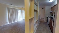 This single-floor space is defined by a notable built-in shelving unit that serves as a partial room divider, separating the carpeted living area from an adjacent tiled space. The shelving structure is finished in a warm yellow tone, offering multiple open shelves ideal for display or storage, and acts as an architectural focal point within the interior.

The living area benefits from generous natural light streaming through large vertical-blind-covered windows, creating a bright and open atmosphere. The carpet flooring in this zone adds warmth underfoot, while the adjoining area transitions to smooth tile flooring, suggesting a functional space such as a dining area or kitchen entry.

A glimpse down the hallway reveals additional rooms beyond, with a floor vent visible along the baseboard, indicating a forced-air heating and cooling system. The walls are painted in neutral tones, complementing the yellow accent of the shelving unit. The overall layout feels practical and functional, with the built-in divider adding a distinctive design element that enhances both the storage capacity and the spatial organization of the interior.