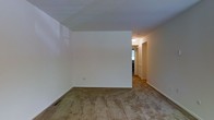 This single-floor space presents a straightforward, functional layout characterized by clean white walls and wall-to-wall carpeting throughout the main living area. The room is open and simply designed, with flat ceilings and neutral tones that provide a blank canvas for personalization. A hallway extends from the main living area, leading to additional rooms and featuring louvered bi-fold closet doors, which offer practical storage access. The flooring transitions consistently through the space, maintaining a cohesive feel. Overhead lighting in the hallway provides illumination to the connecting areas, while a window visible toward the back suggests natural light reaches into the deeper sections of the unit. The overall aesthetic is minimal and utilitarian, with standard baseboards lining the perimeter of the rooms. Floor vents are present, indicating a forced-air climate control system. The space is currently vacant, making it easy to assess the layout and structural details without distraction. While modest in ornamentation, the space offers a functional foundation suitable for a variety of furnishing arrangements and interior design approaches.