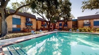 Located at 3737 Hillcroft St in Houston, Texas, this outdoor community amenity space is designed to offer residents a well-rounded recreational experience. At its centerpiece is a generously sized swimming pool with clear turquoise water, framed by a spacious concrete deck lined with blue lounge chairs ideal for relaxing in the Texas sun. Entry to the pool is made convenient with a handrail-equipped staircase, and brick tile accents along the pool edge add a classic finishing touch. Surrounding the pool area, mature oak trees provide generous natural shade, enhancing the comfort of the outdoor environment throughout the day. Adjacent to the pool, a dedicated picnic area features a green table with benches and a bright blue umbrella for additional shade, making it a practical space for outdoor gatherings. A separate children's playground is visible beyond the pool enclosure, equipped with colorful play structures and swings, set beneath the canopy of large trees. The community buildings flanking the amenity area are constructed of warm tan brick with distinctive blue accent details on balconies, shutters, and trim, giving the complex a cohesive and recognizable aesthetic. A gray utility building with a metal roof sits poolside, housing pool-related facilities. The entire area is enclosed by black iron fencing, providing both security and a well-defined boundary for the amenity space.