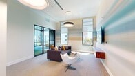 This single-floor space is thoughtfully designed with a contemporary aesthetic, offering a well-appointed living room and a generously sized primary bedroom. The living room serves as the social centerpiece, characterized by elevated ceilings, large windows fitted with roller shades, and abundant natural light that illuminates the neutral-toned carpet flooring. A striking accent wall features a soft, horizontally blended coastal-inspired mural in muted teal, beige, and sand tones, anchored by a wall-mounted flat-screen television above a floating walnut media console. Pendant drum lighting adds a polished, modern touch overhead.

The seating area accommodates multiple lounge configurations, with a dark upholstered sofa, modern swivel accent chair, and minimalist side tables. Black-framed glass partition doors separate the living area from an adjoining room, maintaining an open yet defined layout.

A standout feature of this space is a dedicated golf simulator room, complete with projection technology, an artificial turf surface, and a large-format impact screen. Floor-to-ceiling windows in this area provide outdoor views and additional natural light. The overall ambiance is modern, clean, and amenity-rich, combining comfort with recreational functionality.