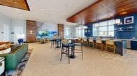 Located at 5440 W Tyson Ave in Tampa, FL, this expansive single-floor amenity space offers a thoughtfully designed community clubhouse with a modern coastal aesthetic. The generous open floor plan accommodates a variety of social and functional zones, all unified by polished terrazzo flooring, soft gray walls, and floor-to-ceiling windows that flood the interiors with natural light and frame views of the surrounding palm-lined landscape.

The main living area serves as the heart of the space, featuring a striking floor-to-ceiling sculptural art installation set against a gray tile accent wall, alongside a sleek linear fireplace and a wall-mounted display screen. Comfortable lounge seating, dining tables, and high-top bar areas are thoughtfully arranged throughout, creating an inviting environment for both casual gatherings and more formal socializing. A wood-paneled ceiling detail adds warmth to the dining zone, which overlooks the outdoors through expansive glazed doors.

The game room includes a billiards table and additional seating, with direct access to an outdoor area framed by palm trees. Private glass-enclosed work suites, labeled with suite numbers and featuring built-in wood desks and textured accent walls, provide quiet spaces for focused work. A wide corridor connects all areas seamlessly, accented with colorful architectural details and abundant recessed lighting throughout.
