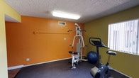 This fitness center is a well-equipped, single-floor exercise space designed to support a range of workout routines. The room is immediately distinguished by its bold color palette, featuring vibrant orange and olive-green accent walls that create an energizing atmosphere. Durable rubberized flooring covers the entire workout area, providing cushioning and traction suitable for exercise.

The space is outfitted with a variety of equipment, including a multi-function cable strength-training machine, a treadmill, an upright stationary bike, and a stability ball, offering options for both cardiovascular and resistance training. A pair of framed wall mirrors are mounted along one wall, allowing users to monitor their form during workouts and visually expand the sense of space. A motivational poster adds to the gym's energetic feel.

Overhead fluorescent lighting ensures the room is well-lit throughout, and a window with vertical blinds brings in natural light, adding to the overall brightness of the area. A textured ceiling runs throughout the space.

An adjacent restroom is accessible directly from the fitness area, finished with wood-look vinyl flooring, a dark-stained vanity with mirror, grab bars, and a clean, functional layout — providing added convenience for users of the facility.