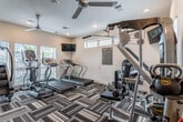 This fitness center is a thoughtfully designed, single-floor facility offering a wide range of equipment to support diverse workout routines. The space is anchored by a prominent Instinct cable machine, complemented by a full rack of dumbbells, kettlebells of varying weights, and medicine balls — providing everything needed for comprehensive strength training. On the cardio side, the facility is outfitted with treadmills, a stair climber, and additional aerobic machines to accommodate a variety of fitness goals. A wall-mounted flat-screen television adds an entertainment element to cardio sessions. Large mirrors line one wall, visually expanding the space and allowing users to monitor their form during workouts. Natural light filters in through multiple windows fitted with blinds, and ceiling fans help maintain a comfortable environment. Recessed lighting throughout ensures the space remains bright and inviting. The flooring features a bold, striped patterned carpet that adds a modern aesthetic contrast to the clean white walls. A frosted-glass door provides access to the facility, and posted fitness center policies ensure a safe and organized environment for all users. Overall, the space balances functionality with a clean, contemporary design.