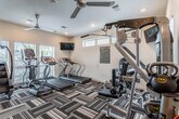 This fitness center is a thoughtfully designed, single-floor facility offering a wide range of equipment to support diverse workout routines. The space is anchored by a prominent Instinct cable machine, complemented by a full rack of dumbbells, kettlebells of varying weights, and medicine balls — providing everything needed for comprehensive strength training. On the cardio side, the facility is outfitted with treadmills, a stair climber, and additional aerobic machines to accommodate a variety of fitness goals. A wall-mounted flat-screen television adds an entertainment element to cardio sessions. Large mirrors line one wall, visually expanding the space and allowing users to monitor their form during workouts. Natural light filters in through multiple windows fitted with blinds, and ceiling fans help maintain a comfortable environment. Recessed lighting throughout ensures the space remains bright and inviting. The flooring features a bold, striped patterned carpet that adds a modern aesthetic contrast to the clean white walls. A frosted-glass door provides access to the facility, and posted fitness center policies ensure a safe and organized environment for all users. Overall, the space balances functionality with a clean, contemporary design.