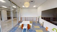 This single-floor children's activity room is thoughtfully designed to foster creativity and engagement in a clean, modern environment. The space is defined by expansive floor-to-ceiling glass walls on two sides, allowing abundant natural light to fill the interior while offering open views of the surrounding outdoor greenery. A sliding glass door provides direct access to adjoining areas, maintaining visual connectivity throughout the facility.

The flooring features a vibrant patchwork carpet in blue, gray, and yellow tones, adding a playful energy to the room. At the center, a child-sized white activity table with coordinating chairs serves as the primary gathering point. Along one wall, built-in natural wood open shelving runs the full length, providing ample storage for materials and supplies at a child-accessible height.

The opposite wall is adorned with charming nature-themed decorative elements, including a bee, star, and woodland animal figures, complemented by a wall-mounted flat-screen TV. A striking globe-style pendant light fixture serves as a focal design element overhead, adding warmth alongside the standard recessed ceiling lighting. The overall ambiance is bright, welcoming, and purposefully designed for young children.
