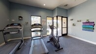 Located at 47750 Adams St in La Quinta, California, this fitness center offers a well-equipped, single-floor exercise facility designed to support a range of workout routines. The interior is finished with cool blue-grey walls, white baseboards, and durable carpeted flooring, creating a clean and functional atmosphere throughout.

The cardio area is equipped with treadmills and an upright stationary bike, positioned near windows that frame views of the surrounding palm-lined outdoor corridor, allowing natural light to fill the space. Adjacent to the cardio section, a designated stretching and functional training area features stability balls and a rack of medicine balls, complemented by standing fans for added comfort during workouts.

Large mirrors line one wall, enhancing the sense of space and allowing for proper form checks. Posted fitness center rules and sauna guidelines are prominently displayed, indicating access to a sauna as an additional amenity. Glass-paneled entry doors open to an outdoor walkway, providing easy access and a seamless connection to the exterior.

Recessed lighting supplements the natural light from the windows, ensuring the space remains bright and welcoming at all hours. Overall, this fitness center provides a practical and comfortable environment for residents or guests seeking a convenient on-site workout facility.