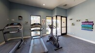 Located at 47750 Adams St in La Quinta, California, this fitness center offers a well-equipped, single-floor exercise facility designed to support a range of workout routines. The interior is finished with cool blue-grey walls, white baseboards, and durable carpeted flooring, creating a clean and functional atmosphere throughout.

The cardio area is equipped with treadmills and an upright stationary bike, positioned near windows that frame views of the surrounding palm-lined outdoor corridor, allowing natural light to fill the space. Adjacent to the cardio section, a designated stretching and functional training area features stability balls and a rack of medicine balls, complemented by standing fans for added comfort during workouts.

Large mirrors line one wall, enhancing the sense of space and allowing for proper form checks. Posted fitness center rules and sauna guidelines are prominently displayed, indicating access to a sauna as an additional amenity. Glass-paneled entry doors open to an outdoor walkway, providing easy access and a seamless connection to the exterior.

Recessed lighting supplements the natural light from the windows, ensuring the space remains bright and welcoming at all hours. Overall, this fitness center provides a practical and comfortable environment for residents or guests seeking a convenient on-site workout facility.