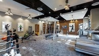 Located at 201 E Gay Street in the heart of West Chester, PA, this single-floor fitness facility combines functionality with a polished, industrial aesthetic. The main workout area is generously sized, offering ample space for both strength training and cardio exercise without feeling cramped. Exposed black ceiling beams contrast elegantly with warm, neutral-toned walls, while a mix of pendant lights and recessed lighting creates a bright, energizing atmosphere throughout. One wall features original exposed brick, adding warmth and character to the space. The gym is outfitted with a comprehensive selection of equipment, including a cable machine, adjustable benches, free weights, and various resistance training stations, catering to a wide range of fitness routines. Patterned carpet tiles cover the floor, providing both comfort and durability underfoot. Mounted flat-screen televisions keep members engaged during workouts, while motivational athletic artwork adorns the walls. Ceiling fans contribute to air circulation throughout the space. Frosted glass-paned doors lead to additional areas within the facility, suggesting private training rooms or supplementary spaces. Overall, the space delivers a well-rounded, professional fitness environment with a distinctive, upscale character.
