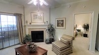 This single-floor space presents a warm and inviting living area defined by timeless design elements and a comfortable, classic aesthetic. The living room serves as the heart of the space, anchored by a traditional white-mantled fireplace with a tile surround, above which a framed artwork adds a refined focal point. Crown molding lines the ceiling throughout, lending an air of architectural elegance to the room.

A ceiling fan with a light fixture provides both practical airflow and ambient lighting, while the soft, neutral wall tones in a muted sage-gray palette create a calm and cohesive atmosphere. Plush carpet underfoot adds warmth and comfort to the space. The room is accessorized with lush indoor plants, contributing a natural, lived-in charm.

Comfortable seating, including a plaid upholstered armchair and ottoman, complements the relaxed yet polished character of the room. A wooden coffee table with a decorative bowl centerpiece grounds the seating area. An open doorway visible from the living room leads to an adjacent bedroom, suggesting a well-connected and functional floor plan that balances openness with defined living areas.
