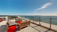This rooftop terrace level offers an exceptional outdoor experience defined by its expansive open-air deck and breathtaking panoramic views of the ocean and surrounding coastline. The outdoor terrace is finished with large-format porcelain tiles and enclosed by sleek glass panel railings, ensuring unobstructed sightlines across the water and beach below. The deck is generously proportioned, accommodating multiple seating arrangements including teak wood benches, lounge chairs, and a full outdoor living set with sofas and accent tables — ideal for relaxation and social gatherings alike.

Access to the terrace is provided through an interior elevator lobby, finished with light wood-look flooring, neutral textured walls, and clean white ceilings. The lobby features stainless steel elevator doors and large floor-to-ceiling windows and glass doors that frame the outdoor terrace and ocean beyond, flooding the interior with natural light. Safety amenities including a recessed fire extinguisher cabinet are integrated discreetly into the lobby walls.

The overall ambiance of this space is bright, airy, and modern, with the interior and exterior seamlessly connected through generous glazing. The combination of refined finishes, open sky, and sweeping coastal views creates an inviting atmosphere that is both functional and architecturally striking.