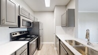 This single-floor residence offers a clean, modern aesthetic throughout, with light wood-look flooring and a neutral color palette that creates a bright, cohesive feel across all rooms. The galley-style kitchen is efficiently designed with gray shaker cabinets, white quartz countertops, a subway tile backsplash, stainless steel appliances including a refrigerator, microwave, and dishwasher, and a stainless steel sink — all illuminated by recessed overhead lighting. The open living area is anchored by a classic fireplace with a white-painted mantle and is framed by crown molding, with sliding glass doors that lead to a private balcony offering outdoor views of surrounding greenery. A hallway connects the living space to the private areas of the home, including bedrooms that feature ceiling fans and additional sliding door balcony access, continuing the bright, airy feel. The bathrooms are finished with large-format gray tile surrounding the soaking tub-shower combinations, paired with gray vanity cabinetry and white countertops. A dedicated laundry area with overhead cabinetry provides added convenience and storage. Throughout the space, consistent finishes and thoughtful design details contribute to a comfortable and functional living environment.