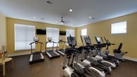 This fitness center offers a dedicated space for cardiovascular training and general exercise. The room is outfitted with a row of treadmills along the windowed wall and a matching row of elliptical cross-trainers, providing a well-rounded selection of cardio equipment for users of varying fitness levels. The machines are modern and feature individual digital display consoles for tracking workout metrics. Multiple wall-mounted flat-screen televisions are positioned throughout the room, allowing users to stay entertained during their workouts. Large windows fitted with horizontal blinds line one wall, inviting generous amounts of natural light into the space and creating an open, airy atmosphere. The walls are painted in a warm yellow tone, contributing to an energizing and welcoming environment. Recessed lighting in the white ceiling supplements the natural light, ensuring the space remains bright at any hour. A ceiling fan provides additional air circulation, enhancing comfort during intense workouts. The flooring features a durable, dark rubberized surface well-suited for a high-traffic fitness environment. Overall, the space is thoughtfully arranged to maximize usability while maintaining a clean, organized layout.
