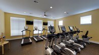This fitness center offers a dedicated space for cardiovascular training and general exercise. The room is outfitted with a row of treadmills along the windowed wall and a matching row of elliptical cross-trainers, providing a well-rounded selection of cardio equipment for users of varying fitness levels. The machines are modern and feature individual digital display consoles for tracking workout metrics. Multiple wall-mounted flat-screen televisions are positioned throughout the room, allowing users to stay entertained during their workouts. Large windows fitted with horizontal blinds line one wall, inviting generous amounts of natural light into the space and creating an open, airy atmosphere. The walls are painted in a warm yellow tone, contributing to an energizing and welcoming environment. Recessed lighting in the white ceiling supplements the natural light, ensuring the space remains bright at any hour. A ceiling fan provides additional air circulation, enhancing comfort during intense workouts. The flooring features a durable, dark rubberized surface well-suited for a high-traffic fitness environment. Overall, the space is thoughtfully arranged to maximize usability while maintaining a clean, organized layout.