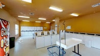 This single-floor laundromat offers a spacious and functional layout designed to accommodate a high volume of laundry needs efficiently. The interior is defined by its warm golden-yellow walls, which give the space a distinctive and recognizable character, complemented by a textured ceiling fitted with rows of fluorescent lighting that ensure the entire area is brightly and evenly illuminated.

The main floor is lined with multiple rows of white top-load washing machines arranged in organized clusters, providing ample capacity for simultaneous use. A generous number of machines are positioned throughout, making this facility well-suited for both individual and high-demand use. Centrally located garment racks and laundry carts offer convenient options for transporting and organizing clothing, while dedicated folding tables provide practical surfaces for sorting and preparing laundry.

Structural support columns are integrated into the floor plan without impeding the flow of the space. The light-colored flooring reflects the overhead lighting, further enhancing the sense of openness. Informational notices are posted on the walls, offering guidance to users. Overall, the space is clean, utilitarian, and thoughtfully organized to create a straightforward and efficient laundry experience.
