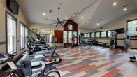 This fitness center is a thoughtfully designed single-level space that combines functional layout with an energizing aesthetic. The architecture is defined by soaring vaulted ceilings that lend an open, airy quality to the interior, complemented by large, dark-framed windows along multiple walls that frame views of surrounding mature trees and allow generous natural light to fill the space throughout the day.

The interior design draws immediate visual interest from the multi-tone plank flooring, which features a bold pattern of red, gray, and natural wood tones arranged in an offset geometric layout. A deep terra cotta accent wall anchors one end of the room, adding warmth and a striking focal point. Large ceiling fans are mounted overhead to maintain comfortable airflow during workouts.

The cardio zone is well-appointed with a row of treadmills, ellipticals, stationary bikes, and rowing machines, positioned to face the windows and wall-mounted televisions. A strength training area offers cable machines, free weights, a weight bench, and a dedicated rack of medicine balls and stability equipment. Floor-to-ceiling mirrors along one wall visually expand the space and aid in form monitoring.

The entryway features glass double doors, wall sconces, and a clean transitional design. A water fountain is conveniently located within the main fitness floor. Overall, the space delivers a polished, motivating environment suited for a comprehensive workout experience.