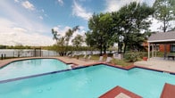 This residential community offers a thoughtfully designed living experience that seamlessly blends indoor comfort with outdoor leisure. The centerpiece of the outdoor amenity area is a generously sized swimming pool with striking blue tile accents, surrounded by a tiled deck furnished with lounge chairs for relaxation. The pool area backs up to a serene waterfront setting, framed by mature trees and enclosed by a wrought-iron fence, providing both privacy and picturesque views of the water beyond.

Inside, the open-concept living and dining area creates a bright, connected space well-suited for everyday living and entertaining. The living room features plush carpeting, ceiling fans with light fixtures, and large sliding glass doors fitted with vertical blinds that invite natural light while offering easy access to the outdoors. A dedicated dining area with hardwood-style flooring sits adjacent to the living space and flows into a functional kitchen, accessible through a distinctive arched passageway.

The kitchen is equipped with dark cabinetry and black appliances, providing a practical workspace. The bedroom continues the comfortable aesthetic with carpeted flooring, soft neutral tones, and coastal-inspired artwork adorning the walls. Throughout the space, a cohesive palette of warm grays and natural wood tones creates a calm and inviting atmosphere.