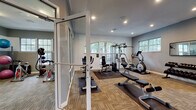 Located at 3021 SR-590 in Clearwater, Florida, this single-level fitness center is thoughtfully designed to accommodate a wide range of workout needs. The facility is organized into distinct exercise zones, each tailored to a specific type of training.

The cardio area features a row of treadmills and an elliptical trainer positioned along a bold teal accent wall, complemented by natural light from arched windows that offer views of the surrounding greenery. A wall-mounted television keeps members entertained during their sessions.

The strength training zone is equipped with a multi-station cable machine, a weight bench, and a stationary bike, all set on rubberized floor mats atop durable carpet tile. Recessed lighting ensures the space is well-lit throughout. A dedicated free weights area includes a full rack of dumbbells and a wall mirror to assist with form and technique.

A separate group exercise and functional training room provides space for cycling bikes, battle ropes, stability balls, and medicine balls. Glass-panel doors connect the rooms, maintaining an open and airy feel throughout the facility.

The facility's interior is finished in neutral grey and white tones, accented by teal walls in select areas. Decorative elements, including a local area map, add a welcoming community touch to the overall environment.