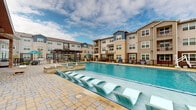 Located at 255 Williamsburg Pkwy in Fate, TX, this resort-style outdoor amenity area is designed to deliver a premium recreational experience. The centerpiece is a generously sized swimming pool with crystal-clear blue water, flanked by rows of in-water lounge chairs that invite residents to relax and unwind. The surrounding paver deck provides ample space for sunbathing, with additional freestanding lounge chairs lining the pool perimeter.

Adjacent to the pool, a covered outdoor patio is attached to the community clubhouse, which features striking stone masonry, large floor-to-ceiling windows, and a warm wood-paneled ceiling. This sheltered seating area is furnished with colorful chairs and tables, creating an inviting social space that opens directly to the pool deck. A pergola-shaded lounge area offers another covered gathering spot, complete with outdoor dining furniture and umbrellas.

The amenity space also includes a dedicated lawn area, ideal for recreational activities, as evidenced by the cornhole boards visible on the deck. A spa or water feature is situated near the pool's edge, adding to the resort ambiance. The multi-story residential buildings surrounding the area feature a mix of neutral siding and blue accent panels with private balconies, completing the cohesive community setting.