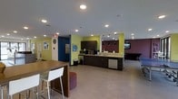 This single-floor communal space is thoughtfully designed to serve as a social and functional hub. The open layout creates a seamless flow between distinct activity zones, making it both practical and inviting. Bold accent walls in lime green, deep blue, and rich plum add a lively, modern character throughout the space, while recessed lighting and pendant lights overhead ensure the area is well-lit at all times.

The kitchenette area is a central highlight, outfitted with dark espresso cabinetry, light stone countertops, a microwave, and a dishwasher — providing all the essentials for preparing and enjoying meals. Adjacent to the kitchenette, a high-top table with white bar stools offers a casual dining or social spot, accompanied by a colorful woven ottoman for additional seating.

A separate lounge area is visible toward the rear of the space, anchored by a wall-mounted television and pendant lighting, creating an ideal setting for relaxation or group gatherings. Light wood-style flooring runs throughout, tying the various zones together with a clean, cohesive look. Wall art and a decorative clock add personal touches that make the space feel welcoming and lived-in. Overall, this communal area balances functionality with an energetic, contemporary aesthetic.