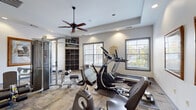 This single-level fitness center is designed to provide a comprehensive workout environment in a clean, well-appointed setting. The space features neutral white walls, recessed lighting, and a ceiling fan that ensures comfortable air circulation throughout the room. Durable, multi-tone carpet tile flooring covers the workout area, offering both practicality and visual appeal.

The facility is outfitted with a diverse selection of exercise equipment, including treadmills, elliptical trainers, a stationary recumbent bike, and a multi-function cable and strength training machine complete with a bench and leg press attachment. Equipment is arranged to allow for ample movement and accessibility between stations.

Natural light filters into the space through multiple windows fitted with horizontal blinds, providing views of surrounding greenery. Framed artwork adorns the walls, adding a welcoming aesthetic to the functional environment. A built-in cubby storage unit offers organized space for personal belongings during workouts.

Interior glass-paneled windows and a glass door connect the fitness center to an adjacent room, enhancing the sense of openness. Fitness center rules are posted on the wall, and recessed spotlights supplement the natural light for consistent illumination. The overall design balances functionality with comfort, creating an inviting space for regular exercise.