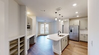 Located at 1735 N Greenville Ave in Richardson, TX, this well-appointed one-bedroom, one-bathroom residence offers a thoughtfully designed single-floor layout that maximizes both functionality and style. The open-concept living and kitchen area serves as the heart of the home, anchored by a central kitchen island with a quartz countertop, undermount sink, and built-in dishwasher. Pendant lighting above the island adds a modern touch, while taupe-toned cabinetry, a subway tile backsplash, and a full suite of stainless steel appliances — including a French door refrigerator, microwave, and range — complete the kitchen. A dedicated bar cabinet with a wine rack provides additional storage and character. Wood-look flooring flows seamlessly throughout the entire unit, lending warmth and visual continuity. The living area benefits from natural light via a glass panel door. The bedroom features multiple windows with blinds, allowing for ample daylight and outdoor views. The bathroom is finished with a long quartz vanity, dual sinks, black hardware accents, a wide framed mirror, and a tiled walk-in shower. A dedicated closet and an in-unit washer connection add everyday convenience. The overall aesthetic is clean, contemporary, and move-in ready.