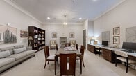Located at 7600 S Mason Rd in Richmond, TX, this single-floor community living room is a thoughtfully designed multipurpose space that balances comfort, function, and refined aesthetics. The room is finished with warm neutral tones, plush wall-to-wall carpeting, and elegant crown molding that frames the ceilings throughout, lending a polished, cohesive feel to the entire space.

A central dining table with upholstered chairs serves as a natural gathering point, lit from above by a striking geometric gold pendant chandelier that adds a modern touch to the classic interior. Flanking this central area are two distinct zones: a cozy library lounge and a dedicated workstation area.

The library lounge features large, dark wood bookshelves stocked with an extensive collection of books, accompanied by a comfortable sofa and warm lamp lighting — creating an inviting nook for quiet reading and relaxation. Framed artwork on the walls adds character and visual interest throughout.

On the opposite side, a long built-in cabinetry workstation with a granite-style countertop accommodates multiple computer stations, making it a practical resource hub. Recessed lighting and an adjoining room visible through a wide doorway further enhance the openness of the space. Overall, this living room exudes a welcoming, community-oriented ambiance with a timeless, well-curated design.