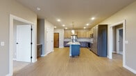 This single-floor residence is designed with a contemporary, open-concept layout that promotes a seamless flow between living and kitchen spaces. The kitchen serves as the centerpiece of the home, showcasing a striking blue island with a quartz countertop, an undermount sink with a matte black faucet, and globe pendant lighting overhead. Cabinetry features a two-tone design, pairing light natural wood upper cabinets with darker lower cabinets accented by black hardware. Stainless steel appliances, including a refrigerator, microwave, and range, complete the functional kitchen. A subtle tile backsplash adds texture throughout. Light-toned luxury vinyl plank flooring extends across the main living areas, contributing to a bright and airy atmosphere. The bedrooms offer plush carpeting, neutral walls, and large multi-pane windows that frame outdoor greenery, filling each room with natural light. A walk-in closet with wire shelving provides ample storage. A dedicated laundry room houses a full-size washer and dryer with additional wire shelving above. The bathroom features a marble-style countertop vanity and clean, neutral finishes, maintaining the home's modern and cohesive aesthetic throughout.