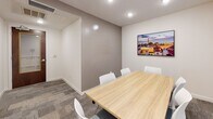 Located on King Street in the heart of Charleston, South Carolina, this single-floor conference room offers a polished and functional meeting environment. The space centers around a light wood rectangular conference table surrounded by contemporary white chairs, providing comfortable seating for a small to mid-sized group. Patterned carpet tile flooring adds texture underfoot while contributing to a professional aesthetic throughout.

Natural light filters into the room through a set of wide windows fitted with horizontal blinds, allowing for adjustable light control. Recessed ceiling lighting ensures consistent illumination regardless of the time of day. A wall-mounted flat-screen display serves as a focal point on one end of the room, suitable for presentations and collaborative screen sharing.

The walls are finished in a neutral palette of white and warm gray, creating a calm and focused atmosphere. Colorful abstract artwork and a cityscape print of Charleston add visual interest and a local sense of place. The room is accessed through a wood-framed glass door, and the layout includes an adjacent alcove area that provides additional flexibility. Climate control is managed via ceiling vents, ensuring a comfortable environment for extended meetings. Overall, the space is well-appointed for professional use in a prime Charleston location.