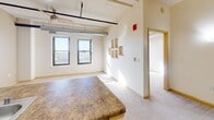 Located at 315 King St in La Crosse, Wisconsin, this single-floor unit showcases a loft-inspired aesthetic with exposed ductwork, visible piping, and track-style ceiling fixtures that reflect the building's industrial character. The open-concept living and kitchen area is anchored by a peninsula counter with a granite-style laminate surface, a double stainless steel sink, and warm wood cabinetry housing white appliances including a refrigerator, range, and built-in microwave. Pendant lighting hangs above the peninsula, complementing the ceiling fan and adding warmth to the space. Neutral-toned carpet flows throughout the main living areas, while large windows allow natural light to fill the room and provide views of the surrounding cityscape. A dedicated utility room includes a stacked washer and dryer alongside built-in wire shelving, offering practical storage. The bathroom features tile flooring, a full-size soaking tub with shower, a vanity with wood cabinetry, and a white countertop. The overall layout, as seen in the floorplan view, is functional and well-proportioned, with clearly defined zones for living, cooking, and daily routines within a cohesive, loft-inspired environment.