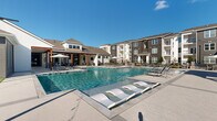 Located at 3445 Bluebonnet Blvd in Baton Rouge, Louisiana, this outdoor amenity complex delivers a resort-inspired lifestyle with a thoughtfully designed array of recreational spaces. At the heart of the property is a generously sized swimming pool with crystal-clear water, surrounded by an expansive pool deck furnished with lounge chairs and in-water sun shelves for relaxing under the Louisiana sun. A covered pavilion with warm wood-slatted detailing provides shaded seating areas adjacent to the pool, offering a comfortable retreat for residents. Complementing the pool area is a well-appointed outdoor kitchen and grilling station paired with dining seating, ideal for gatherings and al fresco meals. The grounds also feature dedicated lawn areas set up for cornhole and other outdoor games, all draped with overhead string lighting for evening ambiance. Lush, professionally maintained landscaping with ornamental grasses, flowering shrubs, and mulched garden beds weaves throughout the courtyards, creating a polished and vibrant atmosphere. A fire pit seating area provides an additional gathering focal point, while the multi-building backdrop, featuring contemporary facades with varied charcoal, navy, and white tones and private balconies, frames the outdoor spaces with modern architectural character throughout the community.