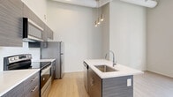 Located at 1021 Euclid Ave in Cleveland's vibrant Midtown corridor, this one-bedroom, one-bathroom unit offers a thoughtfully designed open-concept layout with impressive soaring ceilings that lend a spacious, loft-like atmosphere. The living and kitchen area is anchored by a sleek kitchen island with a white quartz countertop, stainless steel sink, and modern pendant lighting hanging from above. Dark wood-grain cabinetry contrasts beautifully with the bright white walls and light wood-style flooring, which extends throughout the main living area. Stainless steel appliances, including a refrigerator, range, and built-in microwave, complete the kitchen. Exposed ductwork and industrial-style track lighting add a contemporary urban character, while a large black-framed window floods the space with natural light and offers views of the surrounding Cleveland streetscape. The bedroom features soft carpeting, neutral walls, and ample room to furnish comfortably. The full bathroom is finished with large-format tile flooring and a frameless sliding glass shower enclosure, offering a clean and modern aesthetic. A dedicated closet and hallway complete the layout, providing practical storage and easy flow between rooms.