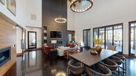 This single-story communal lounge is defined by its dramatic architectural presence, most notably its soaring ceilings that create an expansive, airy atmosphere throughout the open-plan interior. Exposed wooden ceiling beams add a warm, rustic contrast to the otherwise contemporary design, while two large circular chandelier-style light fixtures anchor the space visually and provide ambient illumination.

The focal point of the lounge area is a striking dark feature wall housing a wide linear fireplace, built-in shelving stocked with decorative log arrangements, and a large wall-mounted television with wood-framed detailing. Plush burnt-orange velvet armchairs, a white sofa, and a metallic coffee table create an inviting seating arrangement on a patterned area rug atop polished dark concrete flooring.

Adjacent to the lounge, a substantial dining table with woven barrel chairs accommodates group gatherings. A marble-topped bar counter lined with leather barstools serves a kitchen area featuring a tiled backsplash, open shelving, and stainless steel appliances. A built-in window bench nook provides an additional intimate seating option.

Floor-to-ceiling glass doors along one wall flood the interior with natural light and offer direct access to an outdoor terrace, seamlessly connecting the interior to the exterior surroundings.