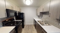 This single-floor unit offers a thoughtfully designed layout that balances functionality and modern style. The galley-style kitchen is a standout feature, showcasing two-tone cabinetry with light upper cabinets and dark lower cabinets, complemented by crisp white countertops and a grey subway tile backsplash. Black appliances — including a gas range, microwave, refrigerator, and dishwasher — provide a bold contrast against the lighter finishes. Wood-look luxury vinyl plank flooring runs throughout the kitchen and living areas, adding warmth and durability. The open area adjacent to the kitchen provides flexible living or dining space, maintaining an airy, uncluttered feel. The bedroom is finished with plush wall-to-wall carpeting and features a ceiling fan with integrated lighting. A window with horizontal blinds brings in natural light and frames an outdoor view of the surrounding landscape. The bathroom is clean and efficiently designed, featuring wood-look tile flooring, a single vanity with mirror, and a full-size soaking tub with a shower curtain rod overhead and grab bars for added safety. The space overall reflects a contemporary, move-in-ready aesthetic with neutral tones and practical finishes throughout.