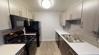 This single-floor unit offers a thoughtfully designed layout that balances functionality and modern style. The galley-style kitchen is a standout feature, showcasing two-tone cabinetry with light upper cabinets and dark lower cabinets, complemented by crisp white countertops and a grey subway tile backsplash. Black appliances — including a gas range, microwave, refrigerator, and dishwasher — provide a bold contrast against the lighter finishes. Wood-look luxury vinyl plank flooring runs throughout the kitchen and living areas, adding warmth and durability. The open area adjacent to the kitchen provides flexible living or dining space, maintaining an airy, uncluttered feel. The bedroom is finished with plush wall-to-wall carpeting and features a ceiling fan with integrated lighting. A window with horizontal blinds brings in natural light and frames an outdoor view of the surrounding landscape. The bathroom is clean and efficiently designed, featuring wood-look tile flooring, a single vanity with mirror, and a full-size soaking tub with a shower curtain rod overhead and grab bars for added safety. The space overall reflects a contemporary, move-in-ready aesthetic with neutral tones and practical finishes throughout.
