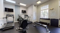 This fitness center in Jacksonville, Florida offers a practical and well-organized workout environment designed to support a variety of exercise routines. The space features durable rubber flooring throughout, providing a safe and comfortable surface for all types of training activities.

The main workout area is equipped with a selection of cardio machines, including a treadmill, stationary cycle, and elliptical trainer, all arranged to allow ample space for movement. A wall-mounted flat-screen television is positioned for easy viewing during cardio sessions. For strength training, a tiered dumbbell rack holds a full range of free weights, while a dedicated medicine ball tower offers multiple weighted balls for functional training. A set of kettlebells is also available, along with an adjustable workout bench for targeted exercises.

Natural light filters in through large multi-pane windows, creating a bright and energizing atmosphere. Recessed and overhead fluorescent lighting ensures the space remains well-lit at all times. The clean, white walls are accented with subtle blue tones, contributing to a fresh and motivating environment. A sanitation station with wipes or spray is conveniently mounted near the entrance, supporting cleanliness and hygiene. Posted fitness rules ensure a respectful and safe experience for all users. The space is neatly maintained and thoughtfully arranged to maximize usability.