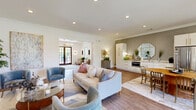 Located at 6300 Moseley Dixon Rd in Macon, Georgia, this thoughtfully designed single-floor space offers a seamless blend of modern elegance and functional comfort. The open-concept layout is anchored by a generously sized living room finished with warm hardwood flooring that flows throughout the entire space.

The living area is bright and inviting, featuring recessed lighting, crisp white walls adorned with crown molding, and large windows dressed with linen drapes that bathe the room in natural light. The seating arrangement includes a plush neutral sofa, tufted blue accent chairs with gold-tone legs, and a round glass coffee table, all centered around a wall-mounted television and a warm wood credenza. A large-scale abstract painting adds a refined artistic touch to the space.

A stylish kitchenette area is neatly integrated along one wall, showcasing floor-to-ceiling white cabinetry with gold hardware, a built-in microwave, a sink, and a textured grey accent wall accented by an octagonal gold-framed mirror. French doors open to the outdoors, allowing natural views to filter into the space and creating an effortless indoor-outdoor connection. The overall ambiance is polished, contemporary, and welcoming — ideal for gatherings or everyday use.