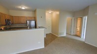 This single-floor unit is designed with an open-concept layout that seamlessly connects the kitchen and living area, creating a cohesive and functional living space. The kitchen is equipped with warm oak-toned cabinetry, a stainless steel refrigerator, and a peninsula with a granite-style countertop that serves as both a workspace and a casual dining or entertaining surface. The flooring transitions from hardwood-style material in the kitchen and entryway areas to plush wall-to-wall carpeting in the living space, providing a comfortable and inviting atmosphere.

The living area is generously proportioned, offering ample room for furniture arrangement. Neutral beige walls and flush-mount ceiling lighting contribute to a clean, understated aesthetic throughout. A floor vent is visible, indicating a central forced-air system for climate control. Multiple doorways are accessible from the main living space — one leading to what appears to be an entry or utility area, and another featuring a bi-fold door, likely enclosing a closet. The overall layout is practical and straightforward, with a neutral palette that provides a versatile backdrop for a variety of interior styles.