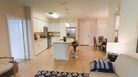 This single-floor space is designed around an open-concept layout that integrates the kitchen, dining, and living areas into one cohesive, flowing environment. The kitchen is a standout feature, showcasing crisp white cabinetry, light granite countertops, and stainless steel appliances including a gas range, built-in microwave, and a French door refrigerator. A central island with a curved edge provides additional counter space and casual seating with modern bar stools, while pendant lighting overhead adds a stylish focal point. Light hardwood-style flooring runs throughout the space, contributing to a warm, unified aesthetic. The kitchen transitions naturally into the dining area, which comfortably accommodates a full dining set, and further connects to the living area, where a patterned area rug anchors the seating zone. Neutral wall tones and recessed lighting maintain a bright, airy atmosphere throughout. The entry door is visible from the main living space, offering a clear sightline that reinforces the open, spacious feel of the layout.