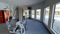 This single-floor fitness center offers a bright and functional workout environment designed to support a well-rounded exercise routine. The space is outfitted with a multi-station cable weight machine that accommodates a variety of strength training exercises, including a leg press and pulley system, making it suitable for full-body workouts. A wall-mounted television is positioned for convenient viewing during exercise sessions. The room is finished with durable, textured carpet flooring well-suited to a fitness environment, while a smooth white ceiling with recessed lighting keeps the atmosphere clean and open. One of the most notable features of this fitness center is its generous use of windows along two walls, flooding the space with natural light and providing clear views of the surrounding outdoor area, including a swimming pool and lush greenery. A decorative potted plant adds a touch of nature to the interior. The light, neutral color palette of white walls and grey flooring contributes to an airy and motivating ambiance. Overall, this fitness center combines practical equipment and a well-lit, inviting atmosphere to create an effective and pleasant space for daily exercise.
