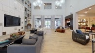 This expansive single-story amenity space is designed to serve as a refined social hub, combining contemporary architecture with carefully curated interiors. The centerpiece of the space is a grand lounge area distinguished by soaring double-height ceilings, oversized globe pendant chandeliers, and light wood-tone flooring laid in a herringbone pattern. Floor-to-ceiling French doors flood the space with natural light and offer views of the surrounding outdoor landscape, while built-in shelving units and a decorative feature wall with inset tile accents and live-edge wood benches add artistic character.

The lounge transitions seamlessly into multiple seating zones, including areas with modular sofas, wingback chairs, and a curated media wall with a mounted flat-screen television framed by dark wood paneling. A second lounge zone features a decorative wall display of framed panels and mirrors, creating intimate conversation areas.

A fully equipped catering kitchen runs along one side of the space, featuring white quartz countertops, white cabinetry, stainless steel appliances, and a wall-mounted television above a white brick accent wall. Adjacent to the kitchen, a café-style dining area offers tufted leather banquette seating, dark circular tables, and woven pendant lighting, with large windows providing ample natural light and garden views throughout.