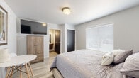 This single-floor unit at 4451 Portrush Rd in Rapid City, South Dakota, offers a clean, contemporary design with a neutral color palette of soft whites and warm grays throughout. The space is thoughtfully laid out, combining sleeping and living areas within an open, efficiently designed floor plan.

The bedroom serves as the central living space, featuring light wood-look flooring that runs throughout the unit, contributing to a bright and cohesive aesthetic. A large window fitted with white horizontal blinds allows for generous natural light while offering privacy control. The room accommodates a queen-sized bed with an upholstered headboard and is equipped with a wall-mounted flat-screen television, a wood-toned armoire, and a compact round dining table with modern chairs — providing functional versatility in a well-organized space. A wall-mounted HVAC unit ensures year-round climate control.

The en-suite bathroom is designed with accessibility in mind, featuring stainless steel grab bars near the toilet, a floating wood-toned vanity with a white countertop, a framed mirror with overhead lighting, and durable wood-look flooring. The overall ambiance of the unit is modern, clean, and practical, making it a comfortable and well-appointed space.