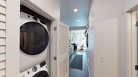 This contemporary single-floor residence is designed with a clean, modern aesthetic that balances style and functionality throughout. The open-concept layout connects the kitchen and living areas seamlessly, creating an efficient and welcoming flow.

The kitchen is a standout feature, outfitted with stainless steel appliances including a range, over-the-range microwave, and dishwasher. Flat-front cabinetry in a muted sage-blue tone is paired with warm wood upper cabinets, while a bold blue ceramic tile backsplash adds texture and visual interest. White quartz countertops provide a crisp contrast and ample workspace.

The living area is bright and airy, with large floor-to-ceiling windows that invite abundant natural light and offer views of the surrounding urban landscape. Dark hardwood-style flooring runs throughout, grounding the space with warmth and continuity. An accent wall in soft sage green adds depth and character to the living room.

A corridor off the main living space leads to a dedicated laundry closet housing a stacked washer and dryer unit, a practical convenience tucked neatly out of sight. The bathroom is finished with a white quartz vanity, undermount sink, large mirror, dark cabinetry hardware, and a glass-enclosed walk-in shower. The overall palette of whites, grays, and muted greens lends the residence a calm, cohesive ambiance throughout.