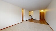 This single-floor unit offers a practical and functional layout with a cohesive, understated design throughout. The open living and dining area is finished with neutral carpet and transitions seamlessly into a hardwood-style floor near the entry and kitchen zone, creating a natural division between spaces. Large windows in the living area allow natural light to flow in while providing views of the surrounding exterior, and a wall-mounted HVAC unit ensures year-round comfort.

The galley-style kitchen is efficiently arranged with white cabinetry, light countertops, and black appliances including a stainless refrigerator and electric range. The hardwood-style flooring continues through this area, contributing to a clean, cohesive aesthetic.

Two bedrooms are each finished with plush neutral carpet and feature windows with blinds that invite natural light. Each room includes a wall-mounted HVAC unit positioned beneath the window, and warm wood-toned trim accents frame the spaces throughout.

The bathroom is accessed through a rich wood-framed door and features a full tub-shower combination with a curtain rod, a wall-mounted sink, white square tile wainscoting, and a classic mosaic tile floor with subtle multicolor accents. The crisp white palette throughout the bathroom gives it a clean, timeless feel.