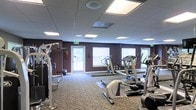 Located at 2904 Calvary Dr in Raleigh, NC, this fitness center offers a dedicated space for exercise and wellness on a single floor. The room features a drop-panel ceiling with recessed fluorescent lighting that ensures bright, even illumination throughout the workout area. Deep plum-colored accent walls give the space a modern, energetic ambiance, while the patterned carpet flooring adds warmth underfoot.

The facility is equipped with a well-rounded selection of exercise machines, including multiple treadmills, an elliptical cross-trainer, a stationary bike, and a multi-function strength training station, providing options for both cardiovascular and resistance training. Large windows fitted with white blinds line the walls, allowing natural light to filter into the space and offering views of the surrounding greenery outside.

Safety is addressed with clearly marked emergency exit signage and a fire extinguisher mounted on the wall. The layout is thoughtfully arranged to allow comfortable movement between equipment. Overall, this fitness center delivers a functional and inviting environment well-suited for a consistent workout routine.