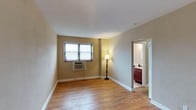 This single-floor space is designed with a clean, functional layout that makes efficient use of its footprint. The main living area is characterized by classic hardwood floors in a warm honey tone, which lend a timeless quality to the interior. Neutral beige walls create a calm, versatile backdrop suitable for a variety of décor styles. A window fitted with horizontal blinds allows natural light to filter into the room, while a wall-mounted air conditioning unit ensures year-round climate comfort. An open doorway leads directly into a bathroom, which features rich, deep-toned cabinetry — likely in a mahogany or cherry finish — adding a touch of warmth and contrast to the otherwise understated palette. The bathroom is well-lit with overhead lighting, providing a bright and practical space. White trim and baseboards throughout lend a crisp, finished appearance to the rooms. The overall ambiance of the space is modest and practical, with straightforward finishes that prioritize comfort and usability. The single-floor layout ensures easy accessibility and seamless flow between the living and bathroom areas.