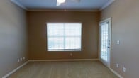 This single-story space features a well-appointed room finished with warm, earthy-toned walls that create an inviting atmosphere. The room is fitted with plush, light-colored carpeting that extends throughout, adding comfort underfoot. Crown molding runs along the ceiling perimeter, lending a refined architectural detail to the space. A ceiling fan is centrally mounted, providing both air circulation and subtle overhead lighting.

Natural light is a defining characteristic of this room, with two windows dressed in horizontal blinds — one standard window centered on the main wall and a larger sliding glass door or French-style window along the adjacent wall. Together, these openings invite an abundance of daylight while offering views of the surrounding outdoor greenery. The blinds allow for flexible light and privacy control throughout the day.

The overall layout is open and unobstructed, making the room versatile and adaptable for various furniture arrangements. White baseboards provide a clean contrast against the warm wall color, further enhancing the neat, well-maintained appearance of the space. The combination of practical features and classic finishes gives this room a comfortable, move-in-ready character.